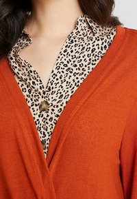 Kvinna med en leopardmönstrad skjorta under en orange stickad cardigan, som visar endast halsen och övre delen av kroppen.