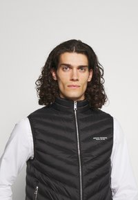 Svart vadderad väst med hög krage, med dragkedja fram och zickzack-stickning. Varumärkeslogotypen visas på vänster sida.