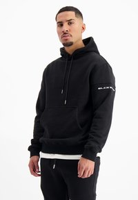 Zwarte hoodie met een voorzak, een capuchon met trekkoord en zilveren accenten. Heeft grafische tekst op de mouw. Zacht materiaal, ontspannen pasvorm.
