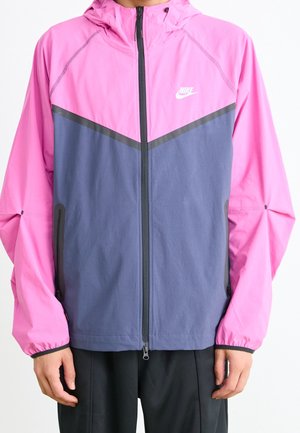 Person, die eine pink-blaue Nike Kapuzenjacke mit Reißverschluss und schwarzen Reißverschluss- und Taschen-Details trägt.