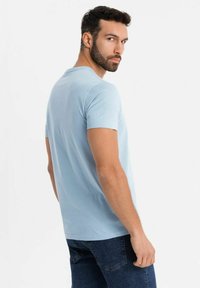 Světle modré bavlněné tričko s kulatým výstřihem, krátkým rukávem a volným střihem, kombinované s tmavými džíny. Textura vypadá měkce a hladce.