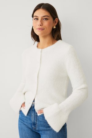 Frau trägt eine weiße, flauschige Strickjacke mit breiten Bündchen und blaue Jeans, steht vor einem einfarbig hellen Hintergrund.