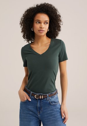 Donna con capelli ricci che indossa una maglietta verde scuro a scollo a V, jeans blu e cintura marrone, in piedi con una mano in tasca.