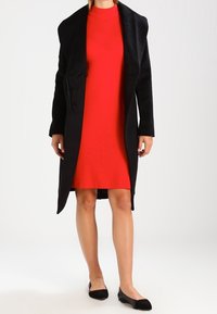 Schwarzer Wollmischmantel mit Schalkragen, offener Vorderseite, über einem taillierten roten gerippten Kleid getragen. Schwarze Ballerinas vervollständigen das Outfit.