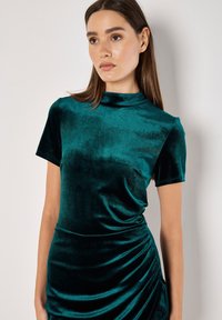 Tiefer Veltkleid mit kurzen Ärmeln und hohem Ausschnitt, das auf einer Seite ein drapiertes Design und eine glatte, weiche Textur aufweist.