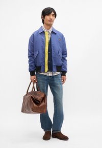 Μπλε bomber μπουφάν με κίτρινη λεπτομέρεια, καρό πουκάμισο, σκούρα ζακέτα, ανοιχτό τζην παντελόνι, καφέ δερμάτινη τσάντα, καφέ μποτάκια μέχρι τον αστράγαλο, μινιμαλιστικός σχεδιασμός.