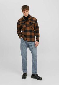 Camisa de franela a cuadros marrón y negra con dos bolsillos en el pecho, cierre de botones, y una capa inferior de manga larga negra en contraste, combinada con unos vaqueros de color azul claro.