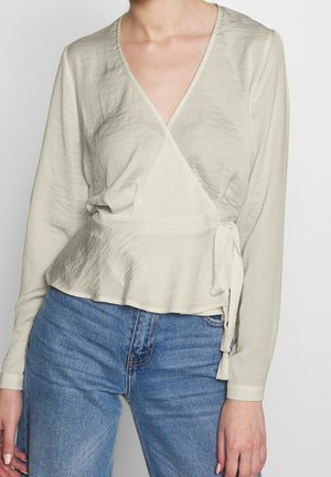 Lys beige omslagbluse med lange ærmer, V-halsudskæring, rynket tekstur og snøre i taljen, parret med blå denimjeans.