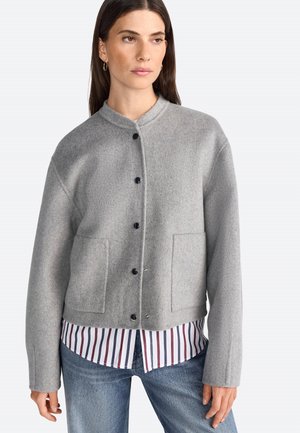 Veste en mélange de laine grise avec un col arrondi, un bouton sur le devant et deux poches avant. Portée sur une chemise à rayures blanche et bleu marine.