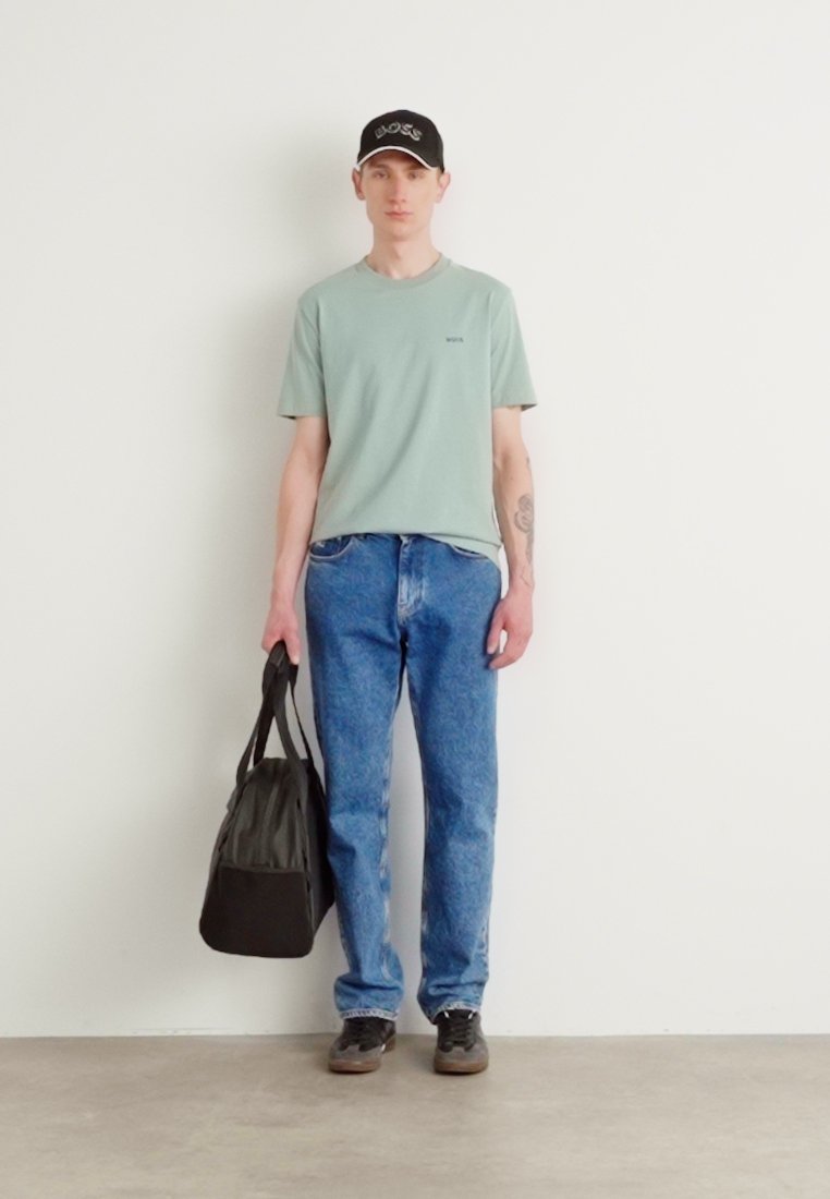 T-shirt à manches courtes vert clair, jeans bleus, casquette de baseball noire, tenant un sac de sport noir, debout contre un mur blanc uni.