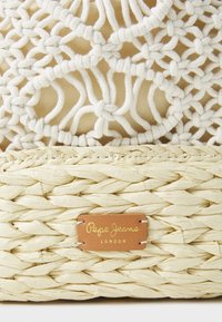 Cestino intrecciato con una base in paglia naturale e un sovraccoperta in macramè bianco di cotone, con una piccola etichetta in pelle impresso "Pepe Jeans London."