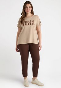 T-shirt beige con la scritta "FRESH" in lettere marroni e frange, abbinato a pantaloni marroni a gamba affusolata e sneaker bianche. Tessuto morbido e casual.