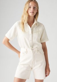 Camicia a maniche corte color crema con bottoni frontali e due tasche sul petto, abbinata a pantaloncini in denim color crema con vita alta e cuciture visibili.