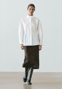 Witte overhemd met knopen en een hoge kraag, met een getailleerde taille; gecombineerd met een bruine, leerachtige kokerrok en zwarte laarzen tot aan de knie.