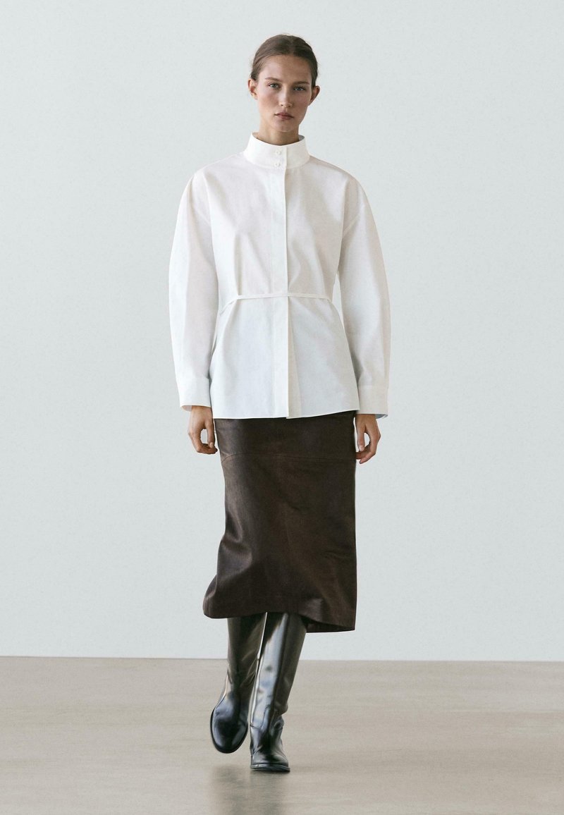 Witte overhemd met knopen en een hoge kraag, met een getailleerde taille; gecombineerd met een bruine, leerachtige kokerrok en zwarte laarzen tot aan de knie.