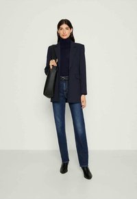 Blazer azul-marinho sobre um suéter de gola alta escuro, combinado com jeans azuis e botas de tornozelo pretas. A modelo segura uma bolsa preta. Design minimalista, ajuste moderno.