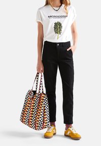 T-shirt bianca con una grafica a foglia verde e testo nero, abbinata a pantaloni neri. Una borsa tote a quadri colorata e scarpe da ginnastica gialle completano il look.