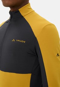 Zwarte en gele lange mouwen fietsshirt met een ritskraag. Heeft een gestructureerde stof en een logo-detail op de borst en bij de mouw.