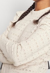 Beige textured blouse with puffed sleeves, featuring eyelet patterns and a round neckline. Fabric has a lightweight, breathable appearance.