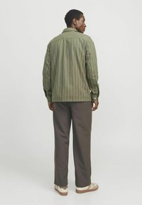Cămașă verde cu dungi, cu mâneci lungi, guler ascuțit și manșete cu nasturi, purtată cu pantaloni maro și adidași de culoare deschisă.
