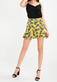 Un haut en satin noir à bretelles associé à une jupe jaune ornée d'un imprimé ananas en vert et marron, avec un ourlet évasé. Des sandales à talons noires.