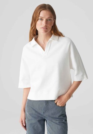 Polo shirt blanc à manches courtes avec un col et une coupe décontractée. Texture lisse, ourlet droit et légères fentes latérales. Assorti à un pantalon rayé.