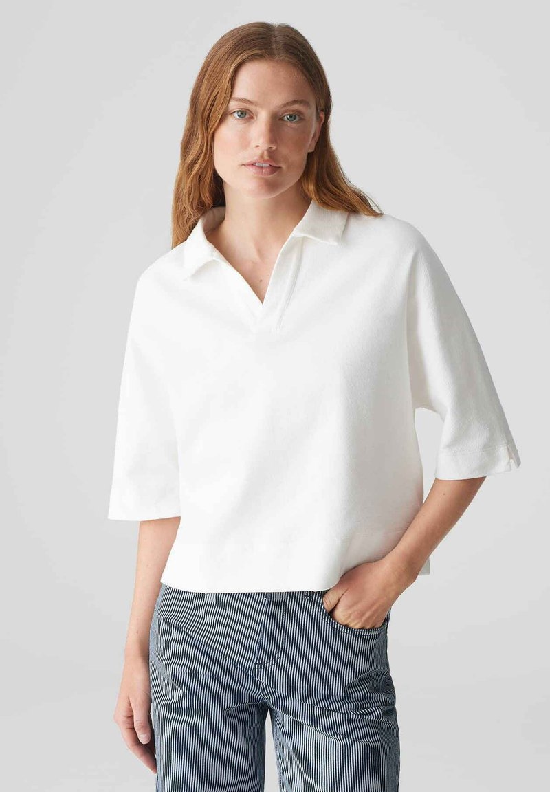 Weißes Poloshirt mit kurzen Ärmeln, einem Kragen und lockerer Passform. Glatte Textur, gerader Saum und leichte Seitenschlitze. Kombiniert mit gestreiften Hosen.