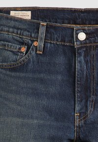 Jean en denim Levi's bleu foncé montrant la poche avant, la passants de ceinture, le bouton et l'étiquette avec les détails de couture.