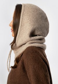 Beige geribbelde capuchon met een gedrapeerd ontwerp, vastgemaakt met trekkoorden, gecombineerd met een bruin gestructureerde trui.