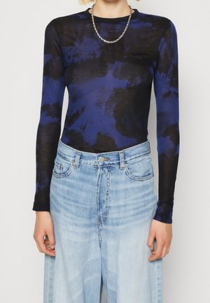 Persona vistiendo una camiseta de manga larga con patrón azul y negro, vaqueros de talle alto azul claro y un collar de cadena plateada.