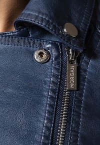 Giubbotto di pelle blu navy con una cerniera argentata, dettagli con bottoni a scatto e cuciture testurizzate. Presenta una linguetta brandizzata sulla cerniera.
