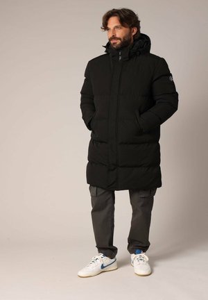 Manteau matelassé noir avec capuche, présentant une fermeture éclair à l'avant et des poches latérales. Associé à un pantalon cargo gris et des baskets blanches avec des accents bleus.