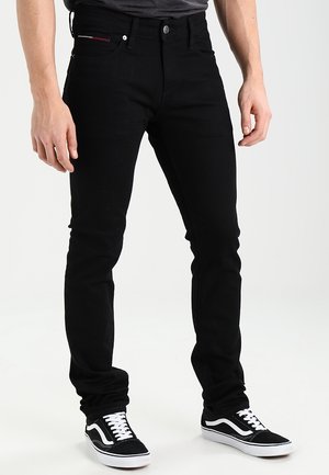 Svart slim-fit jeans med femfickorsdesign, knappstängning och subtila röda sömmar på bakfickan; används med svarta sneakers.