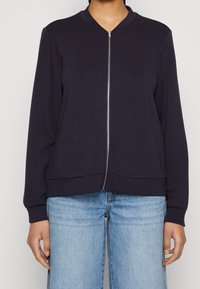 Navy bomber jacket made of soft fabric, featuring a zip front, ribbed cuffs, and two side pockets. Worn over light blue denim jeans.