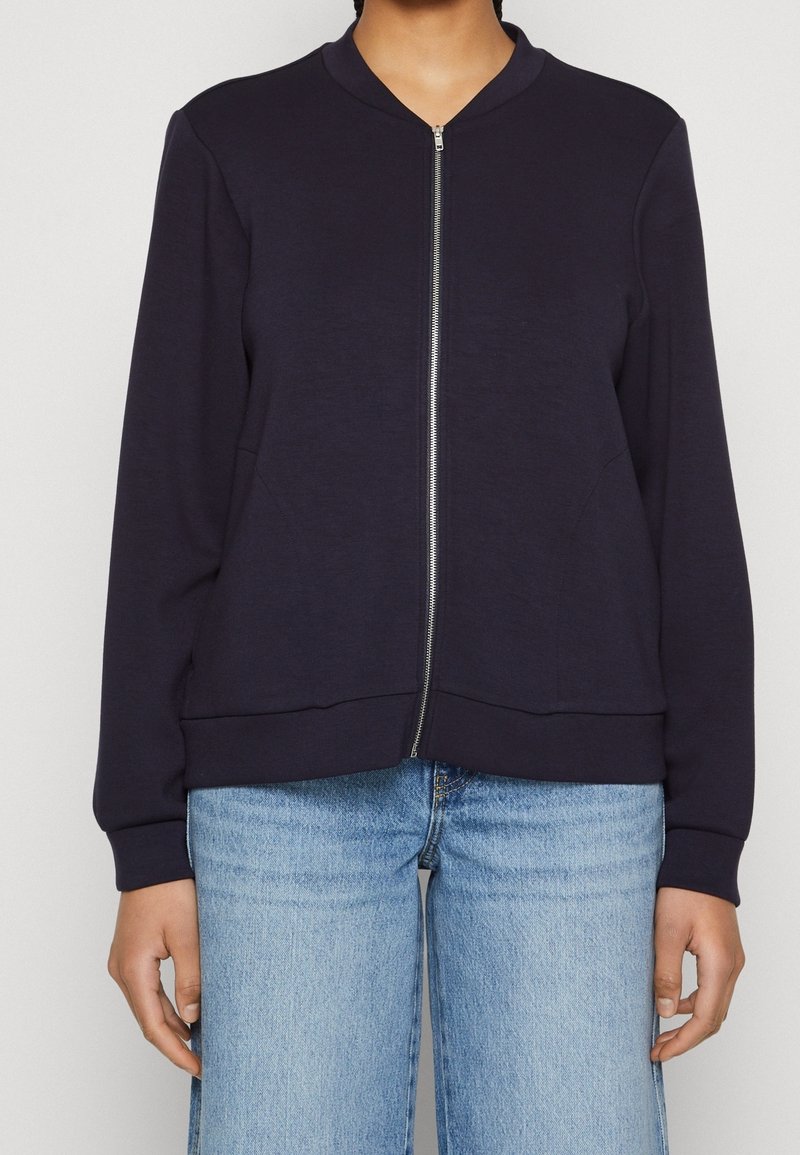 Navy bomber jacket made of soft fabric, featuring a zip front, ribbed cuffs, and two side pockets. Worn over light blue denim jeans.