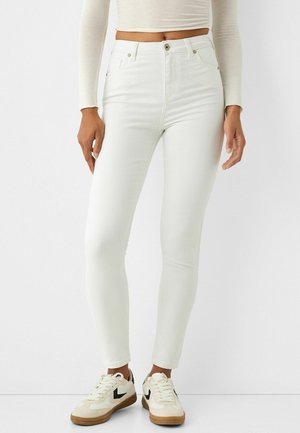 Personne portant un jean skinny blanc taille haute avec fermeture à boutons et des baskets crème avec des touches noires, debout sur un fond blanc.