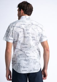 Camisa de manga corta blanca con patrones florales y de paisajes dibujados a mano en negro. Presenta un cuello estándar y un dobladillo curvo.