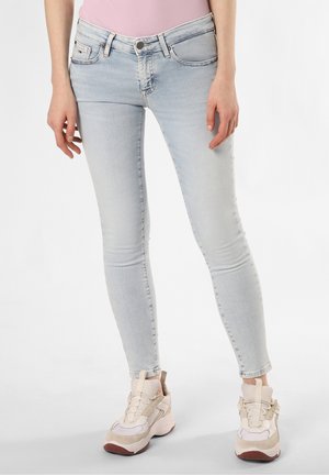 Femme portant un jean skinny bleu clair, des baskets beige et blanches, et un haut rose, debout devant un fond blanc uni.