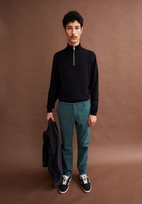 Zwarte trui met hoge kraag, turquoise broek, houdt een donkere spijkerjack vast. Sneakers hebben witte accenten en veters.