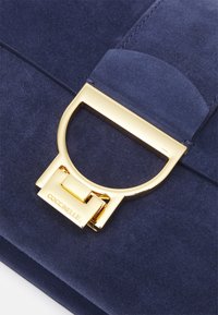 Borsa in suede blu navy con chiusura circolare in tonalità oro. Presenta una trama liscia e cuciture dettagliate. Il nome del marchio "Coccinelle" visibile sulla chiusura.
