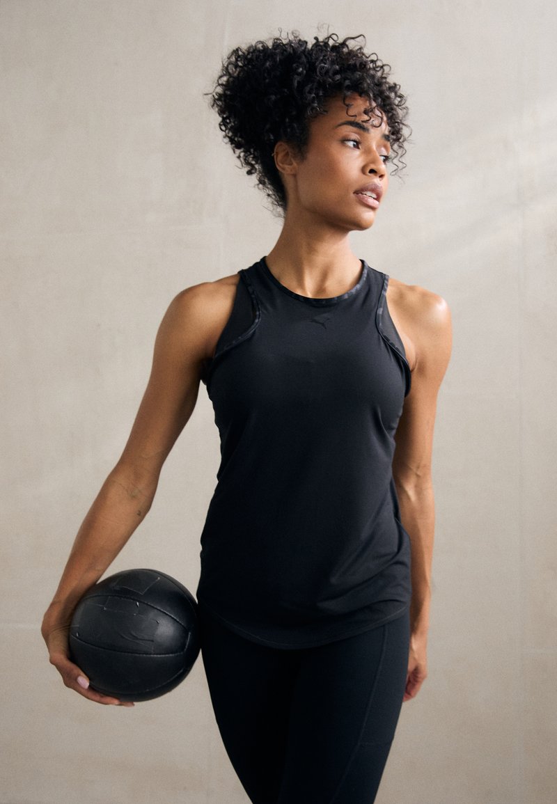 Fekete sportos ujjatlan felső sima textúrával és versenyháts�ó kialakítással, fekete, testhez simuló leggings-szel párosítva. A modell egy fekete medicinlabdát tart a kezében.