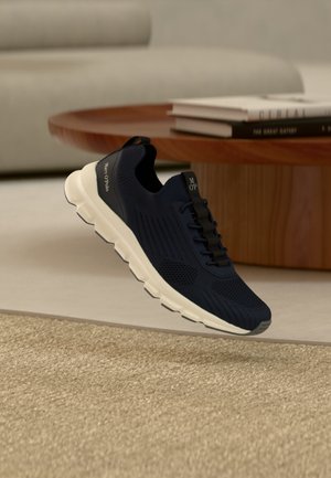 Baskets noires sans lacets avec semelle blanche survolant un tapis beige, à côté d'une table basse en bois avec des livres empilés, dans un salon moderne.