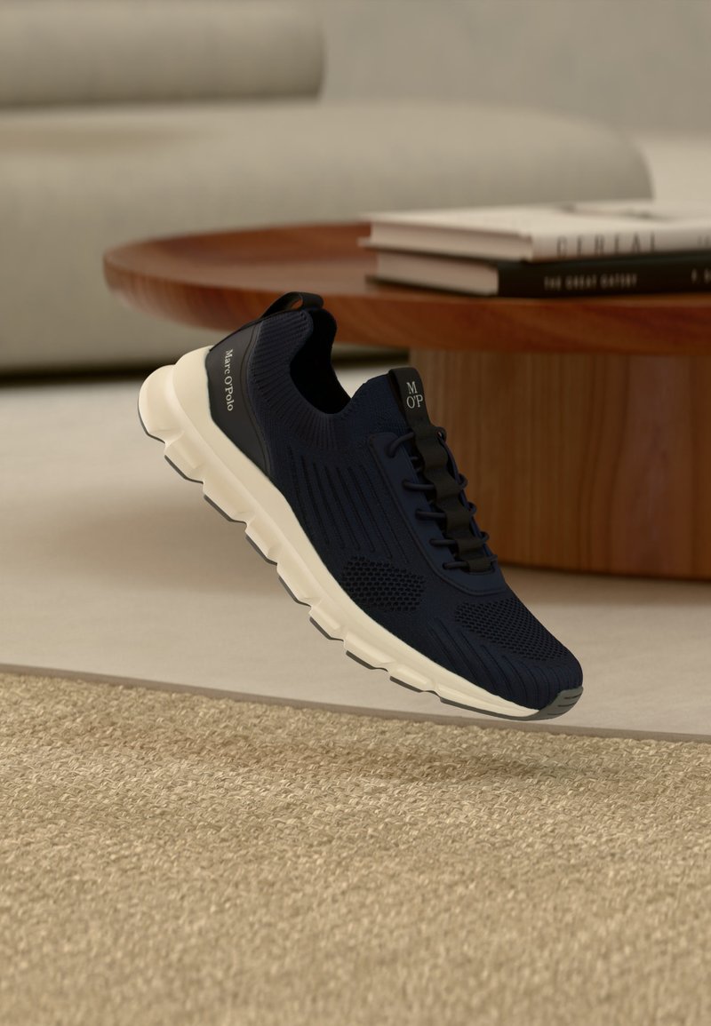 Baskets noires sans lacets avec semelle blanche survolant un tapis beige, à côté d'une table basse en bois avec des livres empilés, dans un salon moderne.