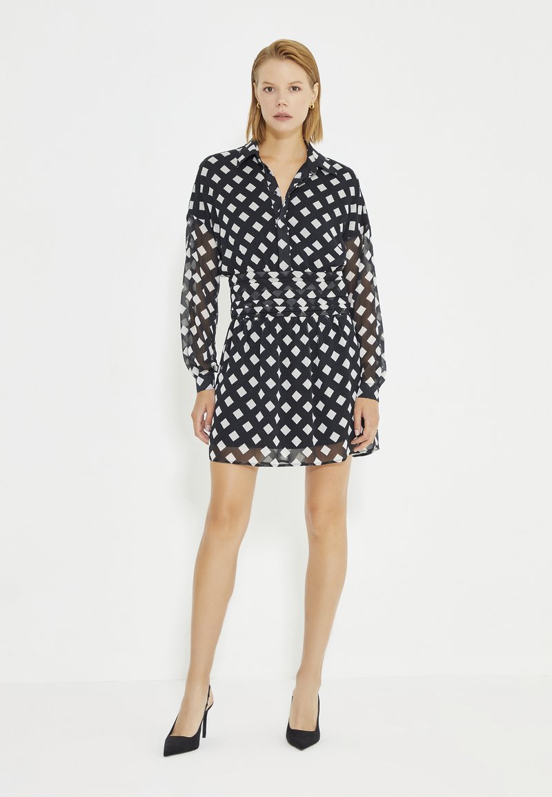 Zwart-wit gestreepte shirtjurk met een kraaglijn, lange doorzichtige mouwen en een geplooide taille, gecombineerd met zwarte hoge hakken.