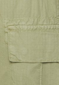 Světle zelená tkanina s mřížkovaným vzorem, obsahující přední kapsu s klopou a prošitými detaily. Hladká textura a kompaktní design.