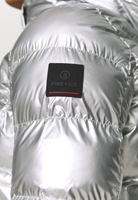 Doudoune argentée avec un motif matelassé et une brillance. Dispose d'une étiquette en tissu noir portant la mention "FIRE+ICE" avec une ligne d'accent rouge.