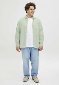 Camisa de botones verde claro sobre una camiseta blanca, combinada con unos vaqueros azules holgados y unos zapatos de lona beige. Fondo neutro.