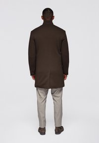 Hnědý vlněný kabát s přizpůsobeným střihem, jedním rozparkem a manžetami na knoflíky, v kombinaci s světle šedými kalhotami a hnědými kotníkovými botami.