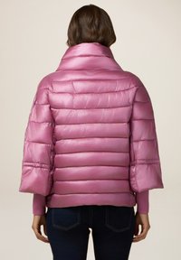 Chaqueta de abrigo rosa con textura brillante y acolchada, cuello ancho, forma rectangular y mangas tres cuartos. Confeccionada en material ligero.