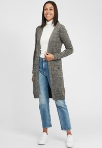 Gray knitted cardigan with a long design. Features side pockets and wooden buttons. Worn over a white turtleneck and paired with blue jeans.